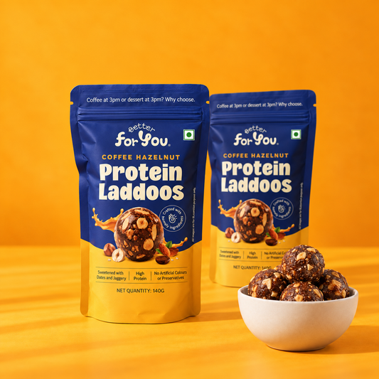 Two stand-up pouches of Better For You Protein Laddoos coffee hazelnut snack are shown on a bright yellow-orange studio background, with a small white bowl of the laddoos in front.