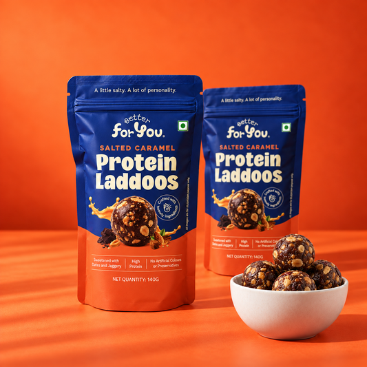 Two blue and orange stand-up pouches of Better For You Salted Caramel Protein Laddoos are displayed on an orange background, with a small white bowl filled with the salted caramel protein laddoos in front.